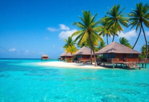 A tropical beach scene featuring overwater bungalows with thatched roofs, surrounded by clear turquoise water. Palm trees line the sandy shore under a bright blue sky with a few scattered clouds.