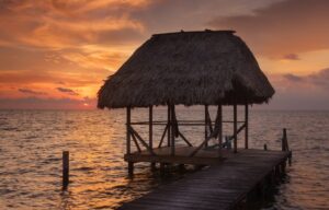 A wooden pier leads to a thatched-roof gazebo over the Belize ocean at sunset. The sky is painted with orange and pink hues, and the water reflects the warm colors. Clouds scatter across the horizon, creating a tranquil scene perfect to relax and rejuvenate in nature's embrace.