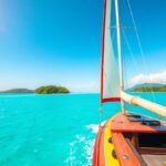 Sailboat gliding through clear turquoise waters under a sunny sky. The boat has a wooden deck and red trim. Small green islands are visible in the distance, framed by a mountainous horizon.