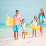 During their family summer vacation in Belize, a family of four strolls along a sandy beach. The father holds a yellow air mattress while the mother carries an inflatable swim ring. The two children clutch yellow toys, both sporting sunglasses. The ocean is calm and the sky is clear.