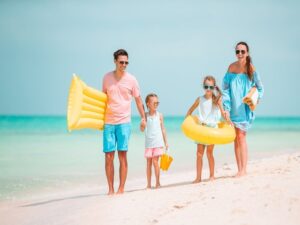 During their family summer vacation in Belize, a family of four strolls along a sandy beach. The father holds a yellow air mattress while the mother carries an inflatable swim ring. The two children clutch yellow toys, both sporting sunglasses. The ocean is calm and the sky is clear.