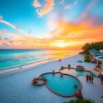 A beachfront scene at sunset with a curvy swimming pool surrounded by people. The sky displays vibrant colors of orange, pink, and blue. The beach is lined with white sand and bordered by ocean waves. Palm trees and buildings are visible on the right.