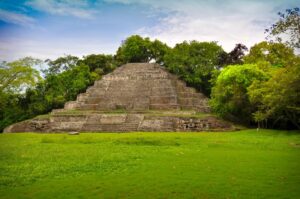 Explore the ancient stone pyramid, a highlight of your Belize itinerary, surrounded by lush green grass and dense trees under a partly cloudy sky. This stunning example of Mesoamerican architecture features several levels with steps leading to the top.