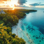 Experience the adventures of ecotourism in Belize with a breathtaking view of a tropical beach at sunset. Turquoise waters, white sandy shores, and lush green forests surround you, while people enjoy the ocean and a boat sails by. A charming building perches on the cliff nearby.