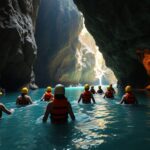 A group of people wearing helmets and life jackets wade through blue water inside a rocky cave, with sunlight streaming in from an opening ahead.