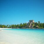 A tropical beach with white sand and clear turquoise water, lined with palm trees. In the background, there is an ancient stone structure resembling a Mayan pyramid under a clear blue sky.