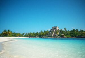 A tropical beach with white sand and clear turquoise water, lined with palm trees. In the background, there is an ancient stone structure resembling a Mayan pyramid under a clear blue sky.