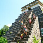 Several children and adults walk down the stone steps of a pyramid-like structure surrounded by greenery, under a clear blue sky. A red informational sign is visible on the right side near the base of the steps.