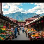 Outdoor market with stalls displaying colorful fruits and vegetables. Several people walk and shop along the central aisle. Buildings and mountains are visible in the background under a blue sky with clouds.