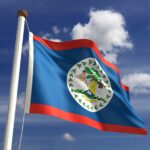 The Belize Flag waves on a flagpole against a partly cloudy blue sky, featuring blue with red stripes and the national coat of arms—a perfect visual for any travel resource or guide to Belize.