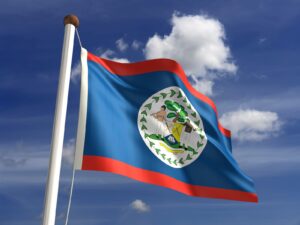 The Belize Flag waves on a flagpole against a partly cloudy blue sky, featuring blue with red stripes and the national coat of arms—a perfect visual for any travel resource or guide to Belize.