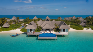 A luxury beachfront villa on stilts at Coco Belize Resort with a private pool overlooks clear turquoise water; nearby, a small boat floats by. Thatched-roof bungalows and lush greenery complete this 2025 Luxury Stays escape under a blue sky.