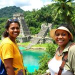 Two women with backpacks smile at the camera in front of ancient stone ruins in Belize, surrounded by tropical greenery and turquoise water, with other people and lush hills completing this geographic guide to adventure.