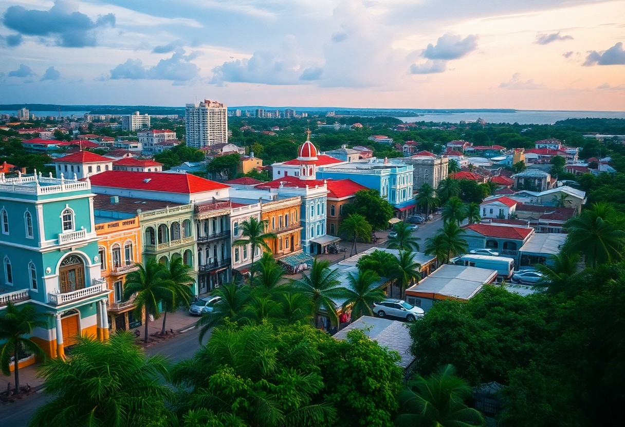 Aerial view of a vibrant coastal cityscape at sunset, showcasing colorful colonial-style buildings with red roofs, lined along a street with palm trees. The background includes greenery, a river, and distant modern buildings under a cloudy sky.