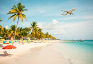 A commercial airplane flies above a tropical beach with white sand, clear blue water, palm trees, and colorful umbrellas. The scene is sunny and a wooden pier extends into the ocean.