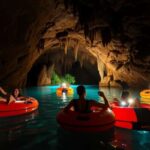 People enjoy an adventure of cave tubing on orange inflatables in a dimly lit cave with striking rock formations. The blue water reflects the beauty of San Ignacio, and green plants glow in the distance under natural light.