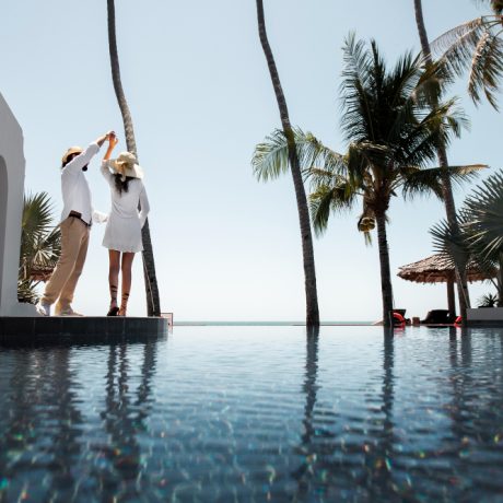 Two individuals enjoying a sunny day by an infinity pool at home with palm trees in the background.