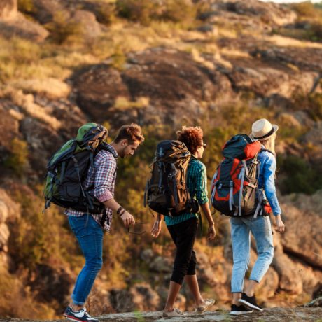 Three hikers with backpacks walking home on rocky terrain at golden hour.